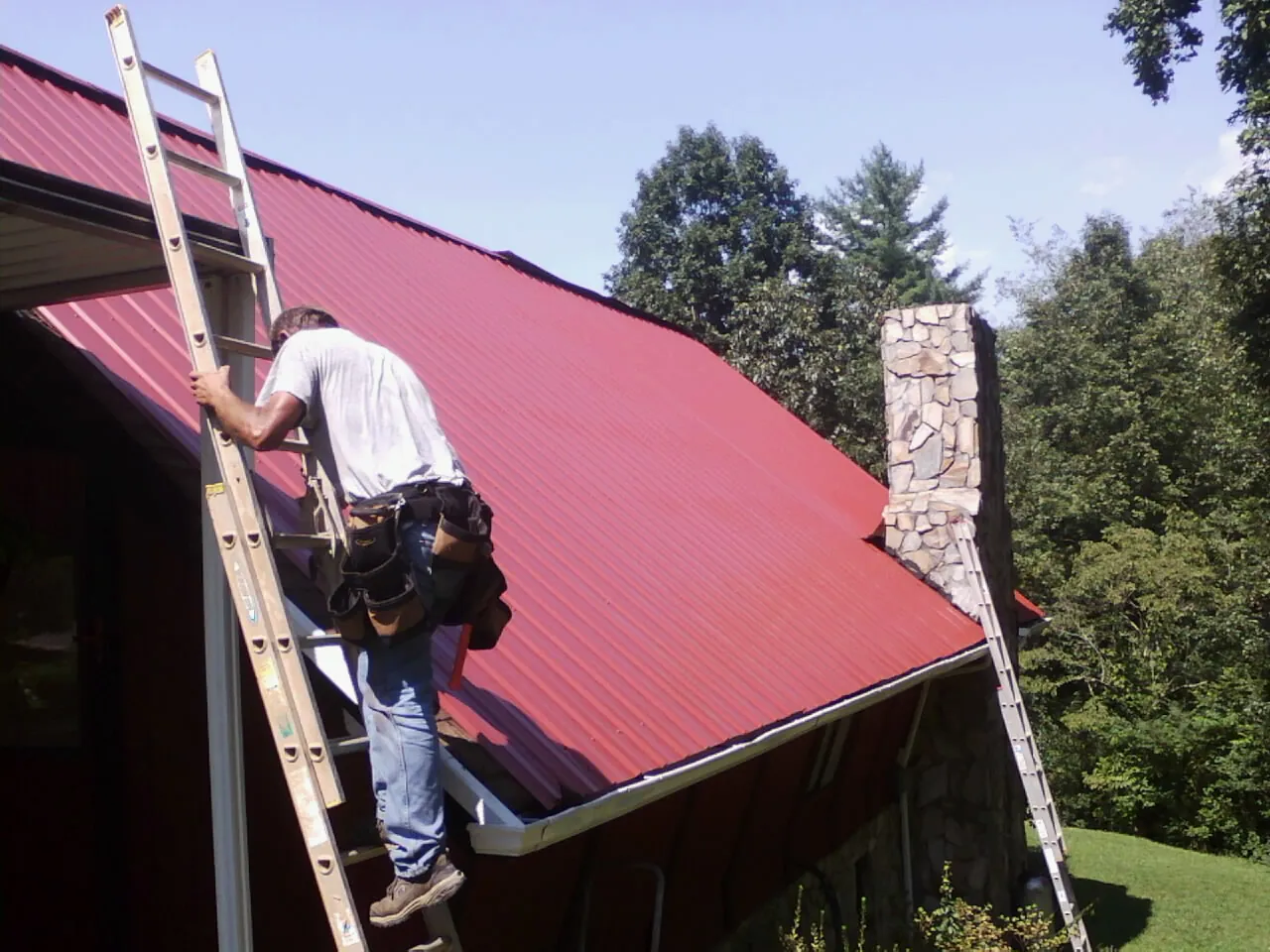 Roofer on a ladder beside a red metal roof and stone chimney — Soffit & Fascia Repair by Roofing Brothers in Attalla